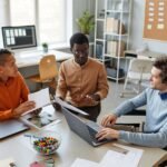 Trois professionnels en réunion dans un bureau moderne, échangeant des idées autour d’un projet sur ordinateur, symbolisant la formation et l’insertion professionnelle en IA.